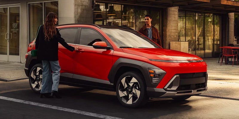 Woman opening the driver's side door of a red Hyundai KONA, with a man standing by the front passenger door.