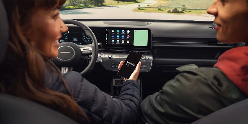Two people in a car; the driver holds a phone with "Android Auto" on screen, which is mirrored on the car's display.