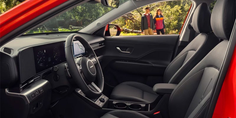 Interior of a red car: dashboard with screen, steering wheel, black seats, and two people outdoors in the background.