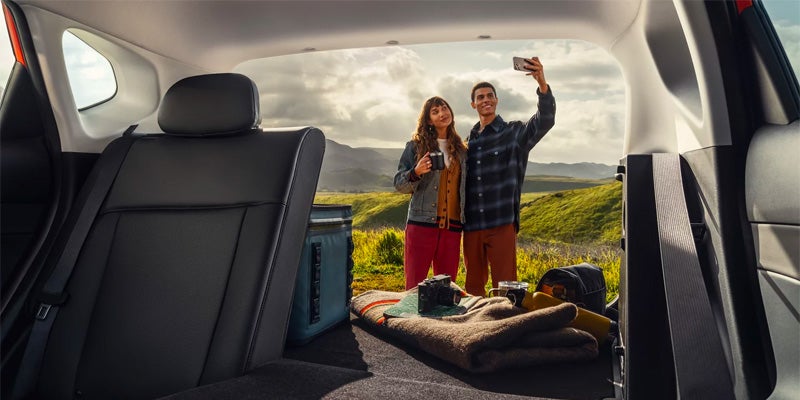View from a car trunk of a young couple taking a selfie with scenic green hills in the background.