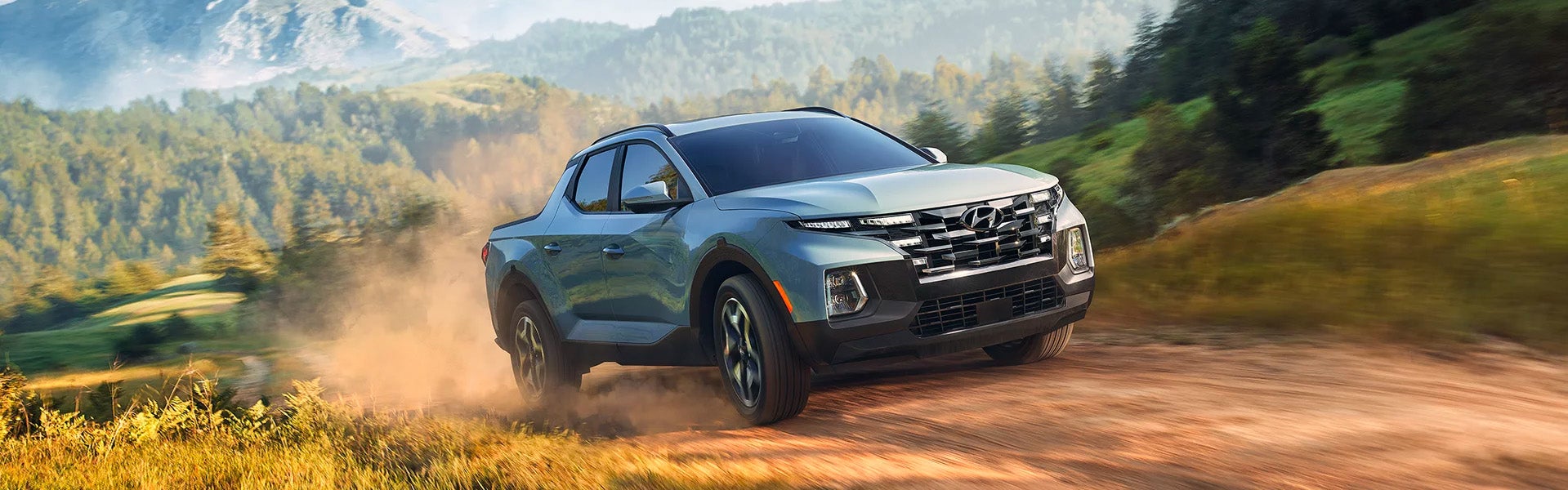 Blue-grey Hyundai Santa Cruz truck kicks up dust while driving quickly on a dirt road through a mountainous forest.
