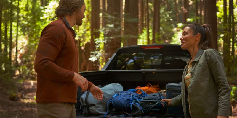Man and woman smiling at each other in a forest with luggage packed in the bed of a pickup truck.