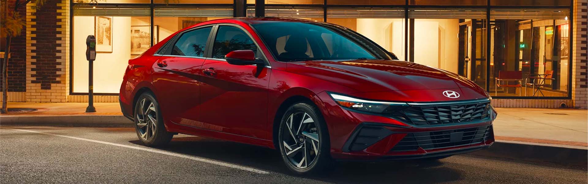 A sleek, dark red Hyundai Elantra sedan parked at dusk, lit by storefronts, on a city street.