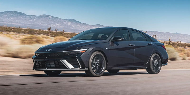 Dark gray sedan with black wheels driving on a desert road with mountains in the background under a blue sky.