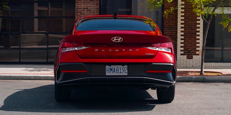 Rear view of a shiny red Hyundai Elantra sedan parked on an asphalt road in front of a brick building.