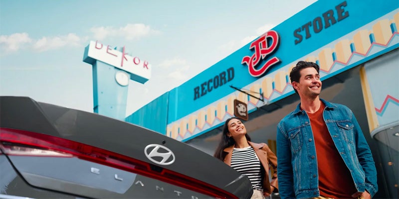 A smiling couple stands in front of a blue record store with a Dekora sign, near a gray Hyundai Elantra trunk.