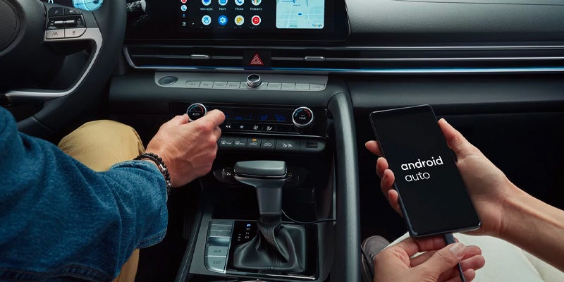 Two people in a car, connected by Android Auto: one adjusting climate controls, another holding a phone showing the logo.