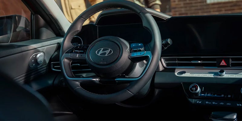 Interior shot of a car dashboard and steering wheel featuring digital screens, analog knobs, and a Hyundai logo.