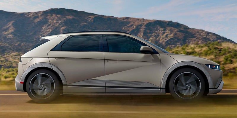 Profile view of a light beige Hyundai Ioniq 5 electric car speeding on a road with mountains in the background.
