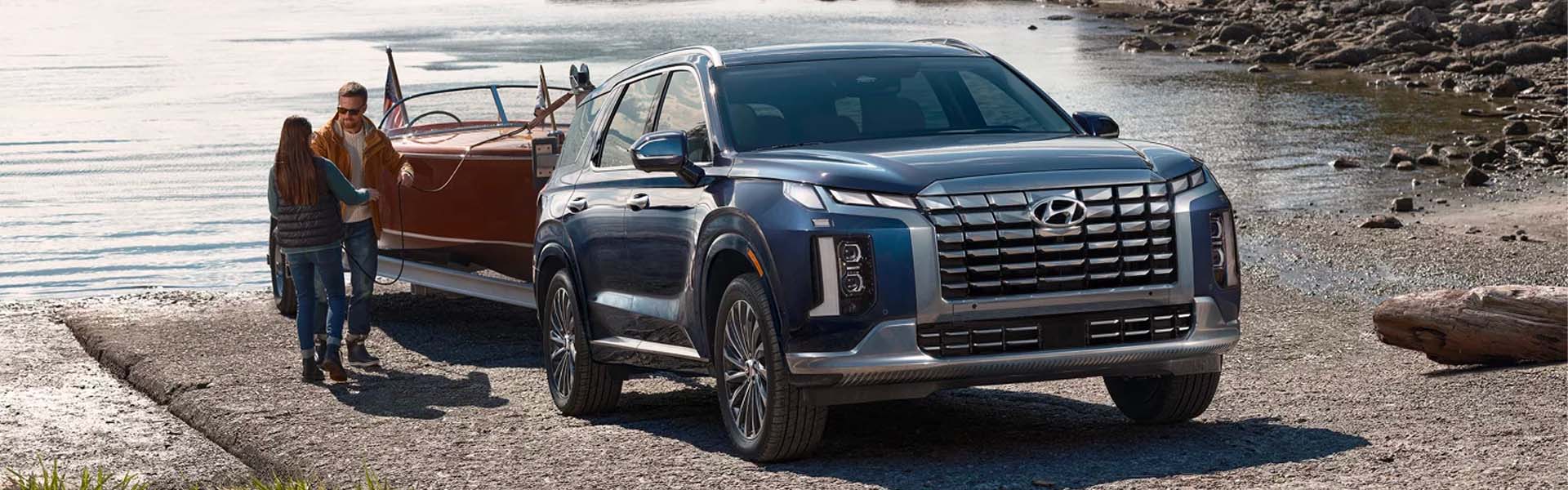 A dark blue Hyundai Palisade SUV tows a vintage wooden boat on a trailer, parked on a gravel ramp next to water.