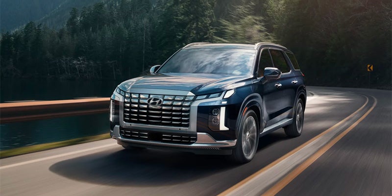 Dark blue Hyundai Palisade SUV driving on a winding road with a forest and lake in the background