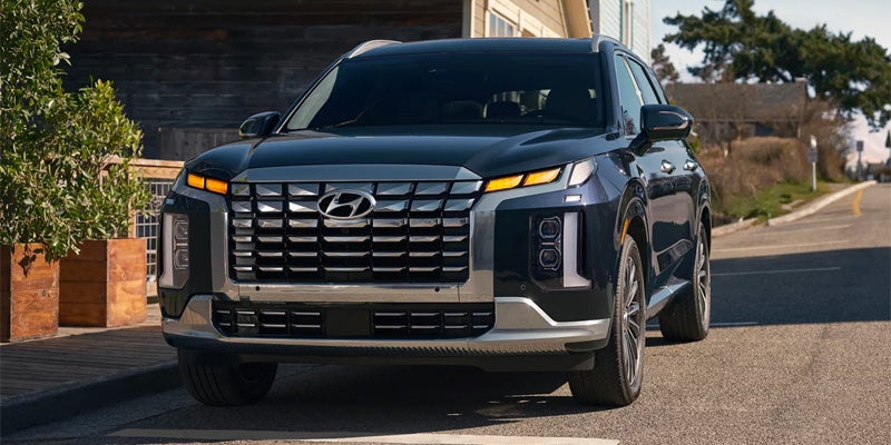 Front view of a dark blue Hyundai Palisade SUV parked on a street with a wooden building and green plants in the background.