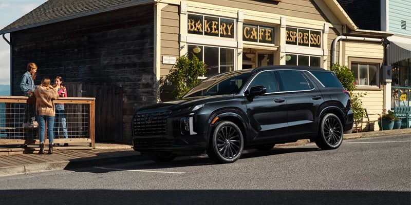 A black SUV parked on the street in front of a bakery and cafe, with three people on a porch to the left.