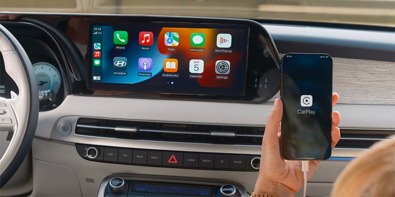 A person's hand holding a phone with the CarPlay logo, connected via USB to a car dashboard with a CarPlay screen.