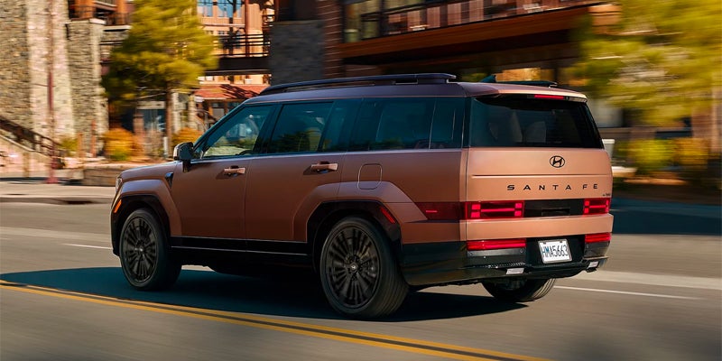 Rear view of a bronze 2024 Hyundai Santa Fe SUV driving on a city street with buildings and trees.