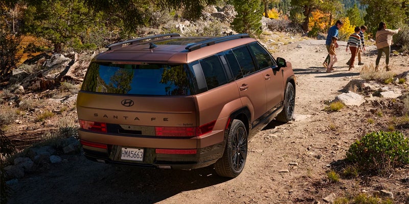 A bronze Hyundai Santa Fe hybrid parked on a dirt path in a forest, with a family exploring nearby.