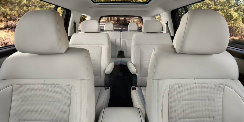 Interior shot of a car, showing three rows of light gray leather seats with a sunroof and trees visible outside.