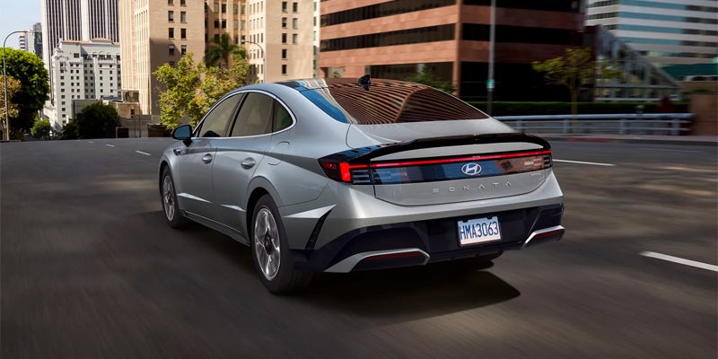 Rear view of a silver Hyundai Sonata driving on a city street, with buildings and trees in the background.