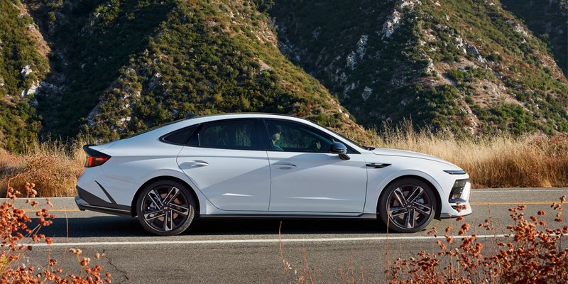 White sports sedan paused on a mountain road with sunlit hills and dry brush in the background.