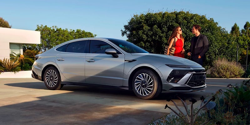 Stylish silver sedan on a modernist driveway with a couple standing by it and lush landscaping.