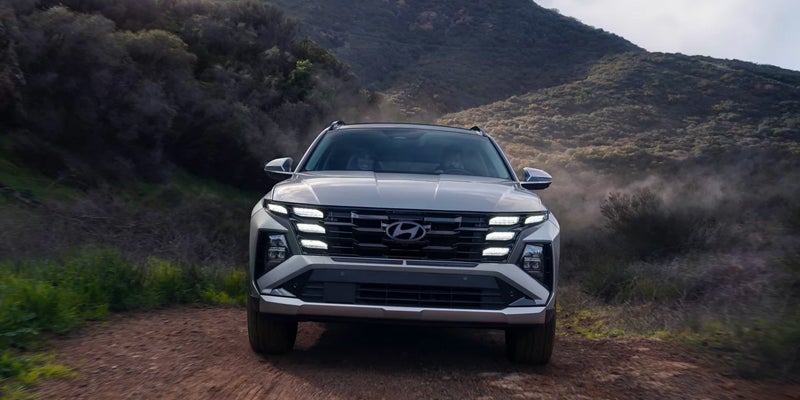 A silver Hyundai Tucson SUV drives on a dusty dirt road towards the viewer, surrounded by brush-covered hills.
