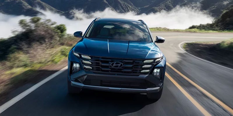 Front view of a dark blue Hyundai Tucson SUV driving on a winding mountain road with hazy peaks in the background.