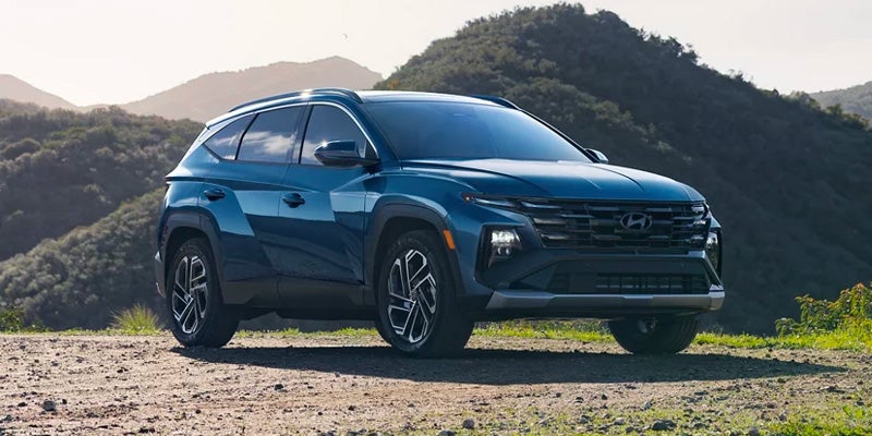 A dark blue Hyundai Tucson SUV parked on a dirt road, with green, tree-covered mountains in the background.