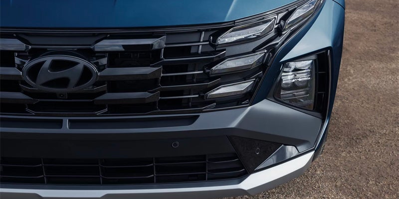 Close-up of a blue SUV's front grille, featuring a dark Hyundai logo, intricate lighting, and lower bumper.