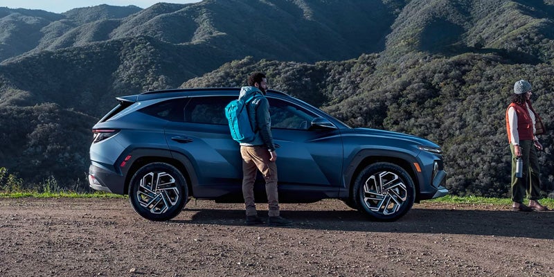 Two hikers with backpacks stand by a blue SUV on a dirt road, with mountains in the background.