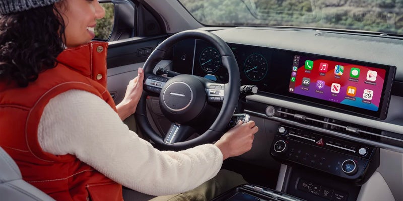 A driver, wearing a red vest over a white sweater, holds the steering wheel and car keys inside a modern car.