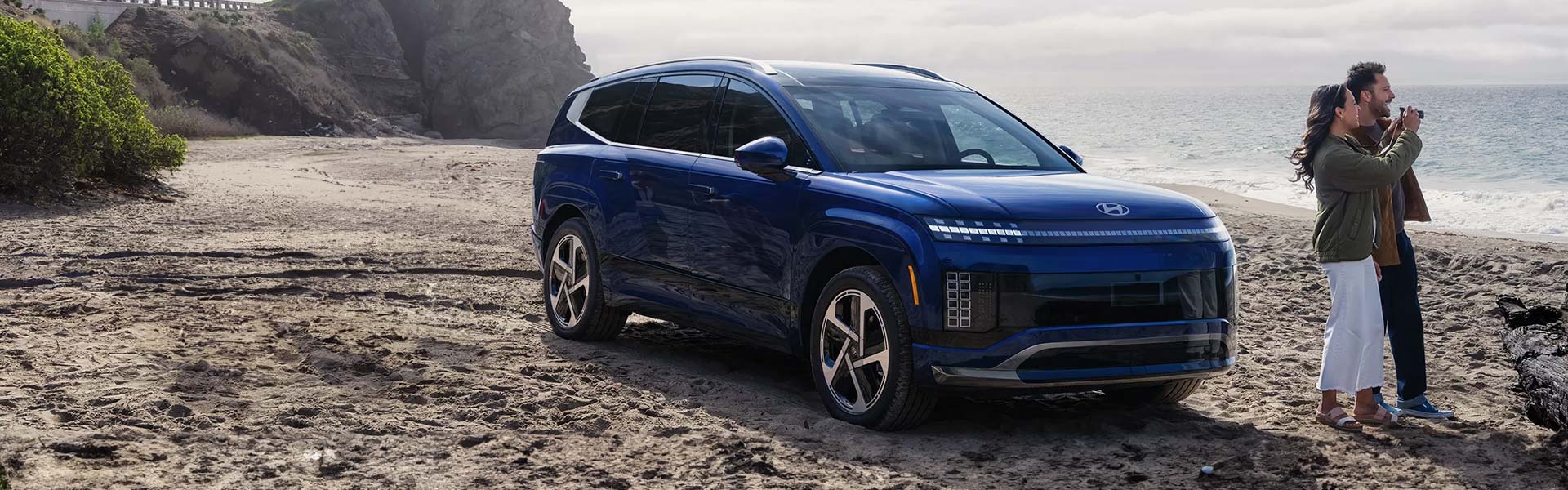 A blue Hyundai IONIQ 7 SUV parked on a sandy beach next to the ocean, with two people looking out at the water.
