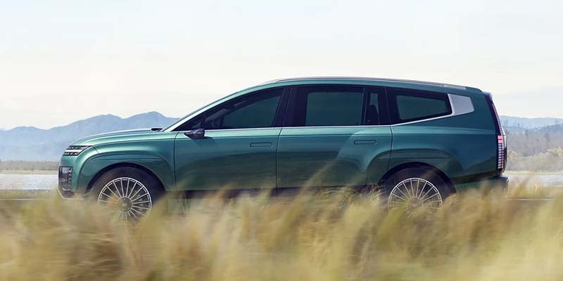 Side profile of a dark teal SUV with a boxy rear, parked in tall light-brown grass with mountains in the background.