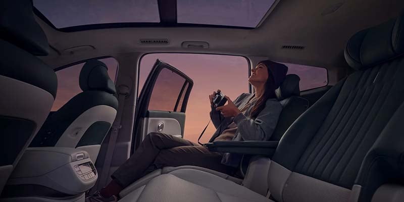 A woman with a camera sits in a car with reclined seats, looking up through the panoramic sunroof at a sunset sky.