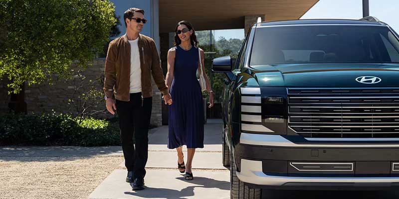 Smiling couple, dressed in brown and blue, holding hands while walking past a dark green Hyundai Palisade.