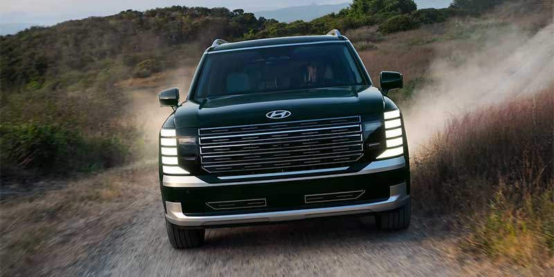 Front view of a dark green Hyundai Palisade SUV driving on a dusty, unpaved road with hills in the background.