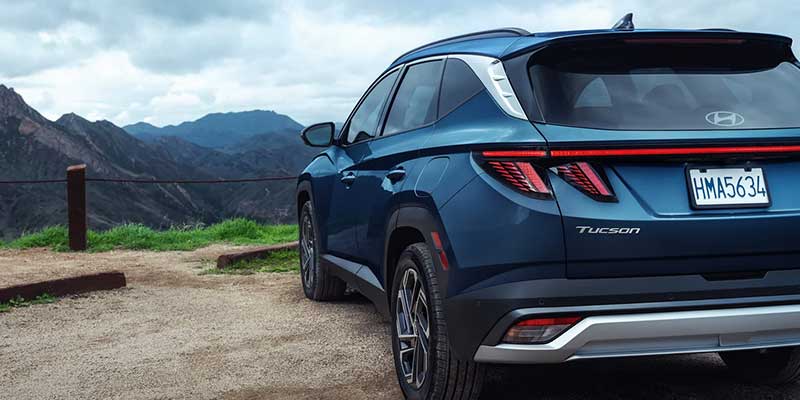 Rear exterior view of a blue Hyundai Tucson SUV parked on a dirt lot overlooking a mountainous landscape.