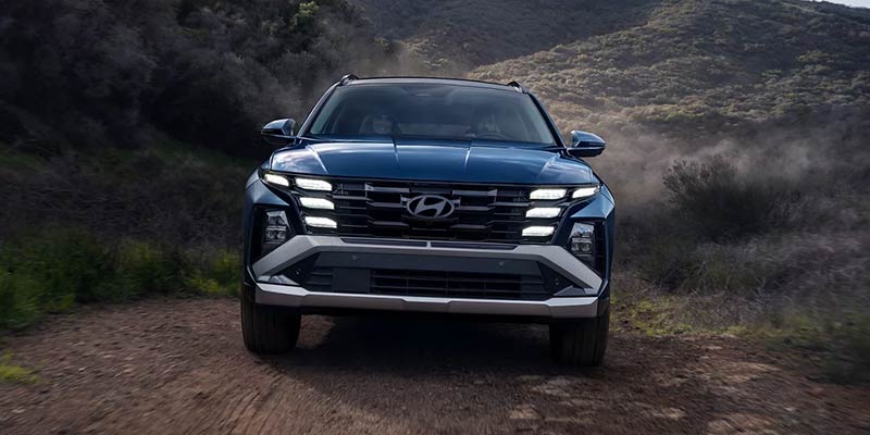 A dark blue Hyundai Tucson SUV drives head-on toward the viewer on a dirt road, amidst dusty, dark green hills.