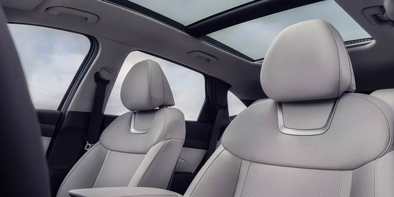 The front two seats of a car with light gray leather and textured fabric, and an open sunroof.