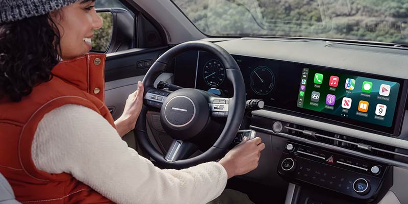 A person in a beanie and red vest sitting in a car, hands on the steering wheel, with car controls and navigation screen visible.